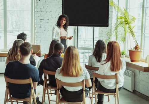 Female african-american speaker giving presentation in hall at workshop. Audience or conference hall. Rear view of participants in audience. Conference event, training. Education, diversity, inclusive concept.